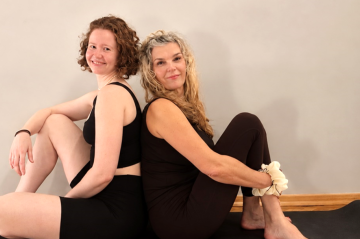 Die beiden Yogalehrerinnen sitzen Rücken an Rücken auf einer Yogamatte.